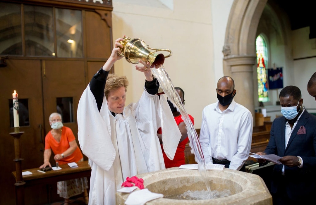 Rev'd Edward pours water into the baptismal font while others look on.